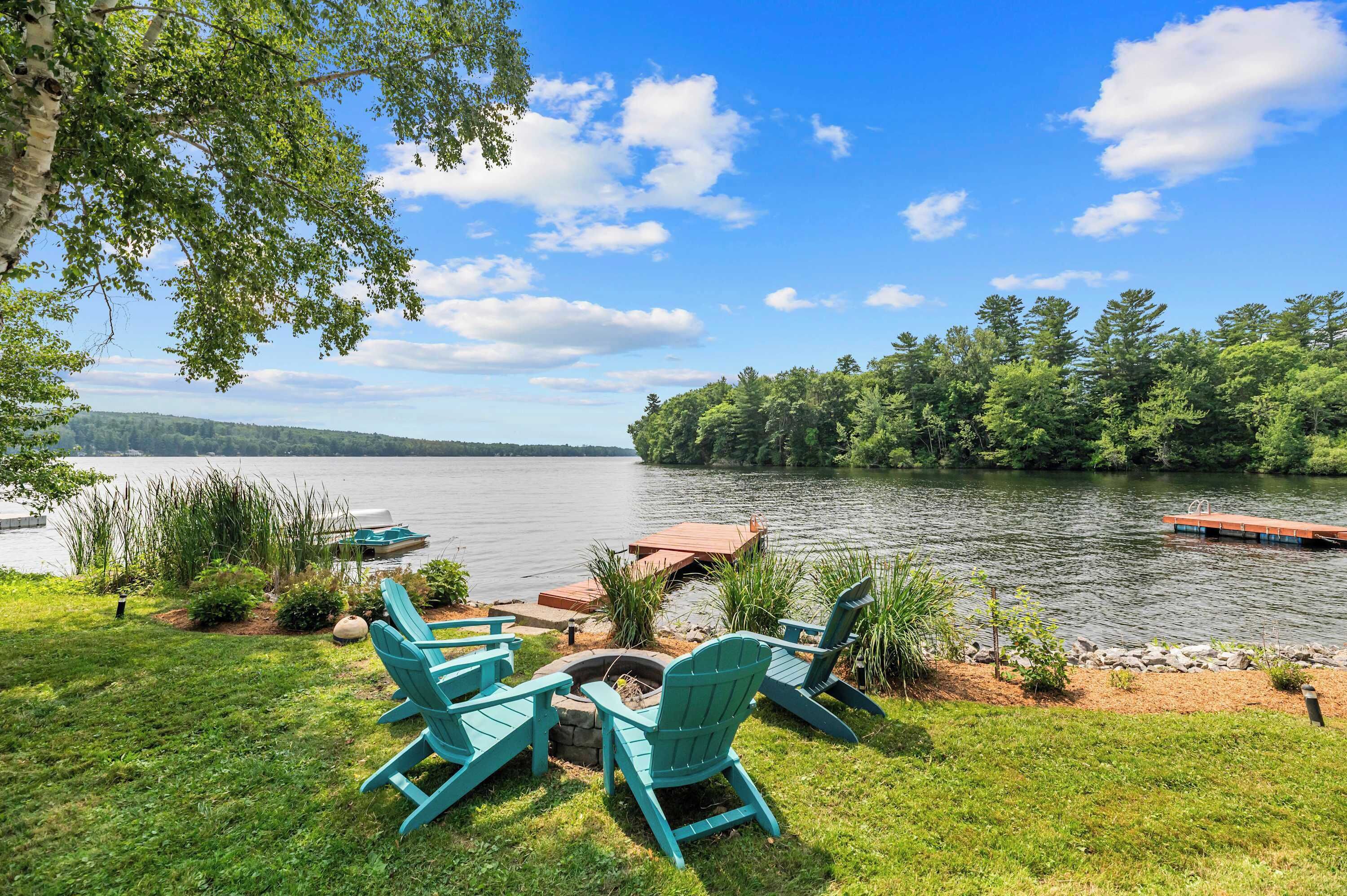 A lakeside view with a dock, chairs, and a fire pit.