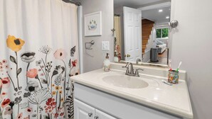 Une salle de bain avec un lavabo blanc, un miroir, un rideau de douche à motifs floraux et un tableau encadré au mur.