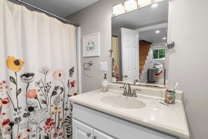 A bathroom with a white sink, a mirror, a shower curtain with floral patterns, and a framed picture on the wall. - Little Landing Cottage | Lakefront · Dock · Kayaks (Winthrop)