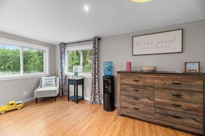 A room with a wooden floor, a desk with a lamp, a water dispenser, a chair, and a dresser with drawers.