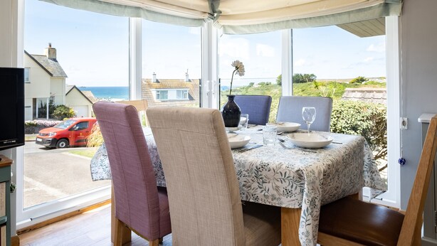 A dining area with a table set for two, large windows with curtains, and a view of the sea and houses.