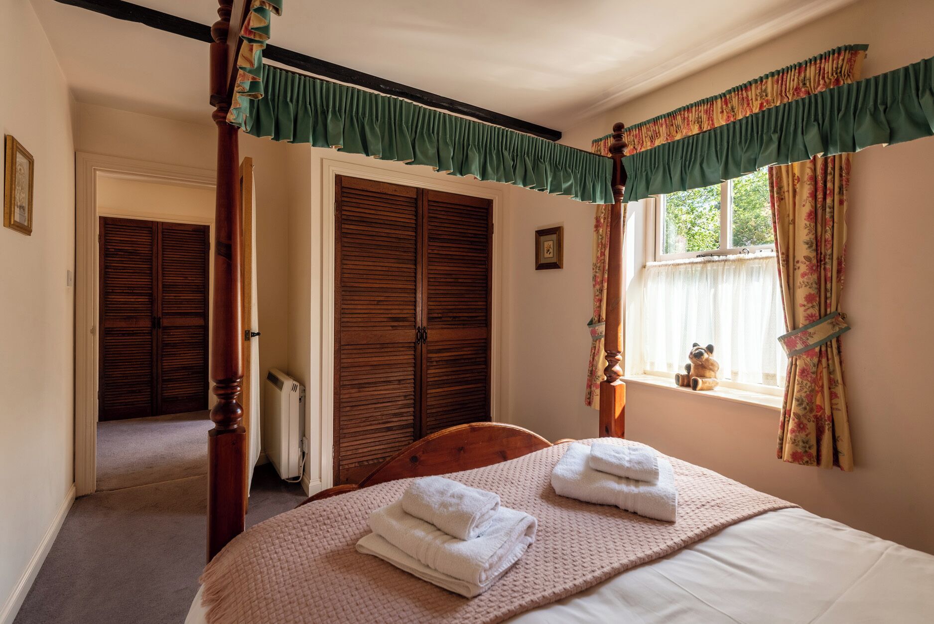 A bedroom with a four-poster bed, a window with floral curtains, and a view of the outdoors.