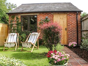 Ein kleines Backsteinhäuschen mit einer Holztür, zwei gestreiften Liegestühlen und einem Garten mit rosa Blüten.