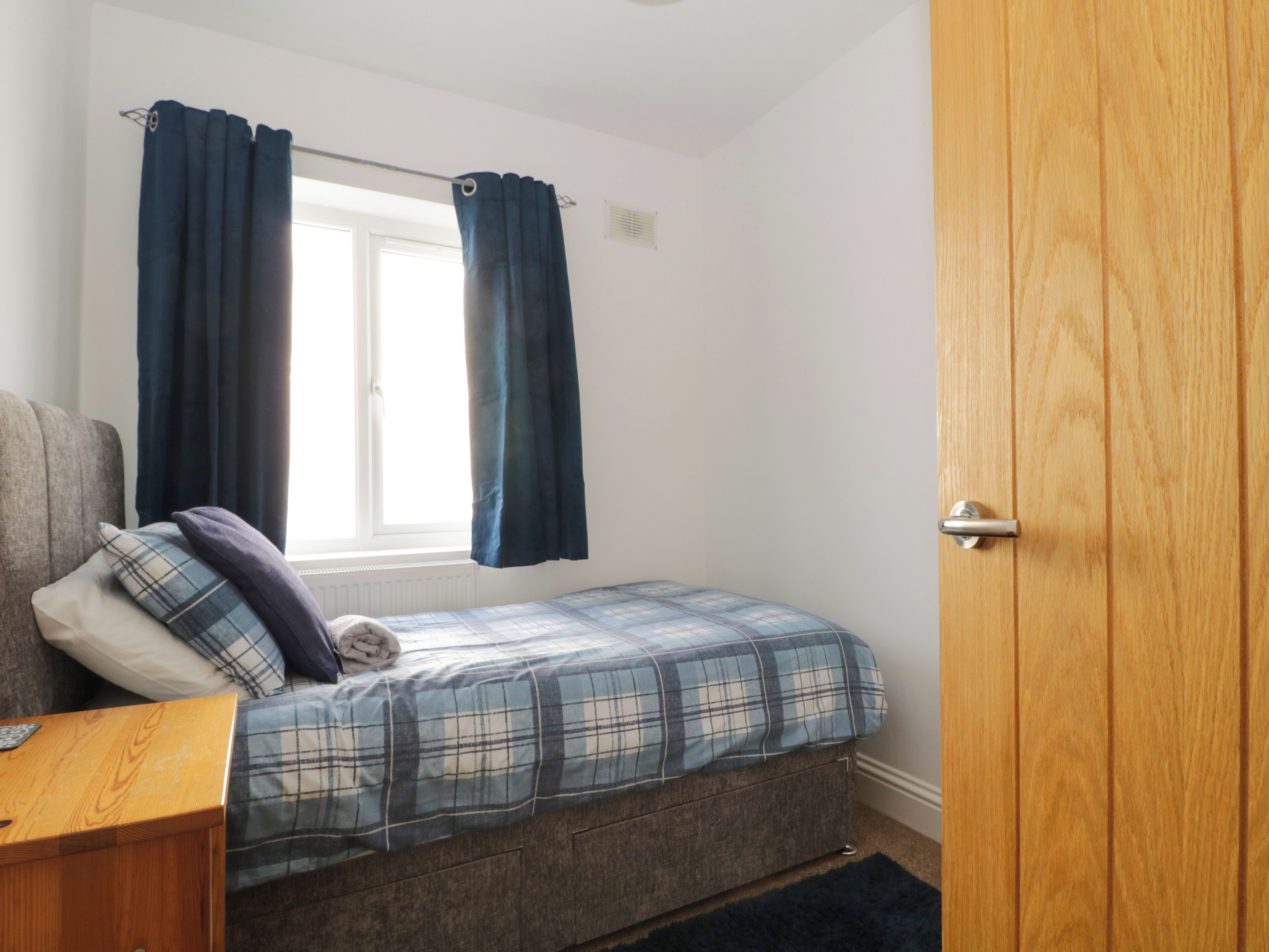 A bedroom with a bed, a window with curtains, a wooden door, and a small wooden table.