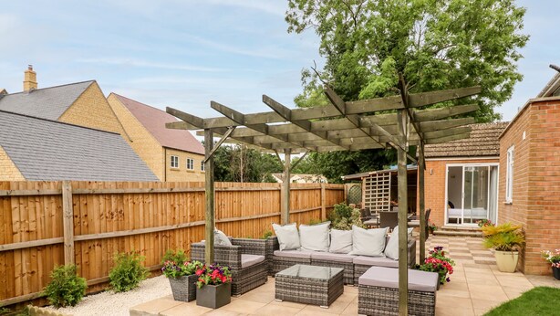 Un patio con mobiliario de mimbre, un pérgola y vista a una casa al fondo.