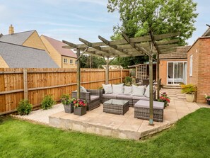 Un patio con mobiliario de mimbre, un pérgola y vista a una casa al fondo.