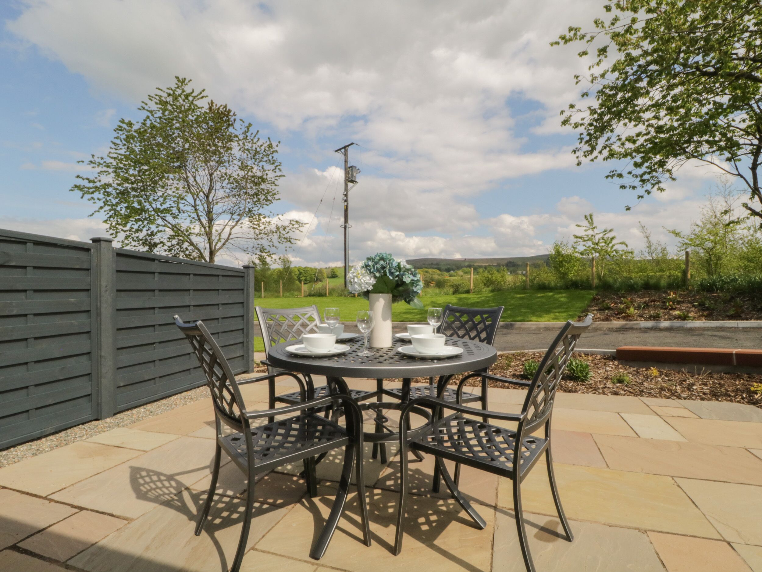 A patio with a round table and four chairs, a vase with flowers, and a view of a green landscape.