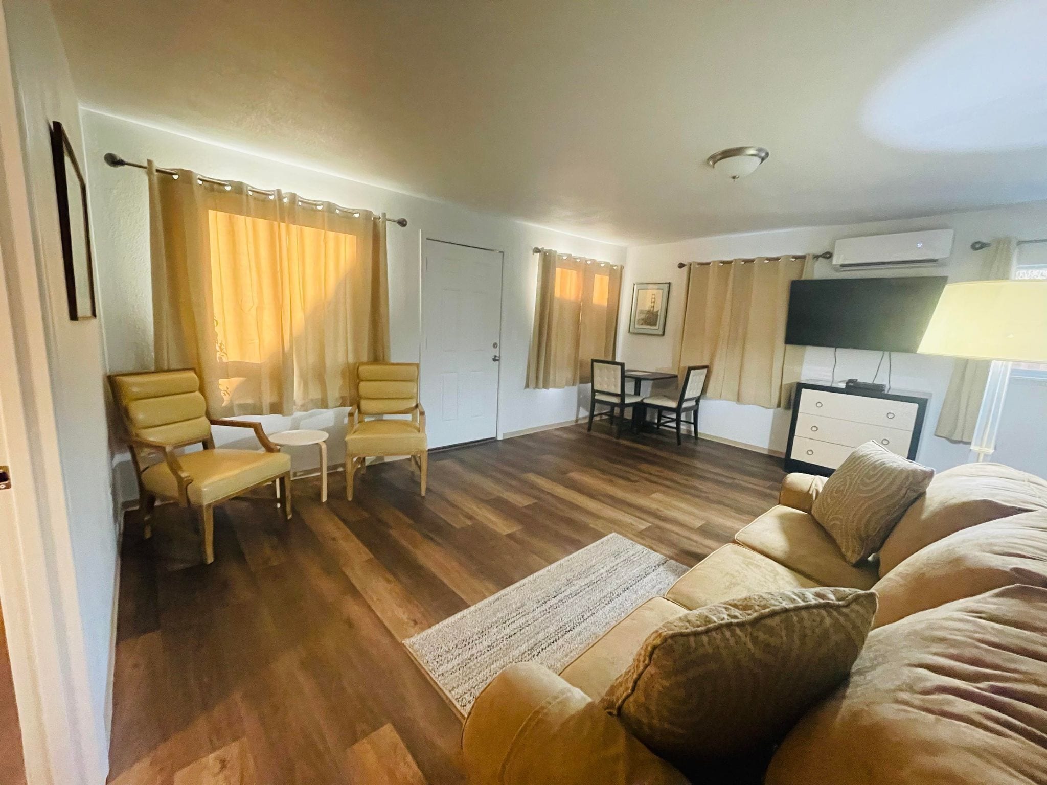 A room with a wooden floor, a sofa, two chairs, a small table, a TV, and an air conditioning unit.
