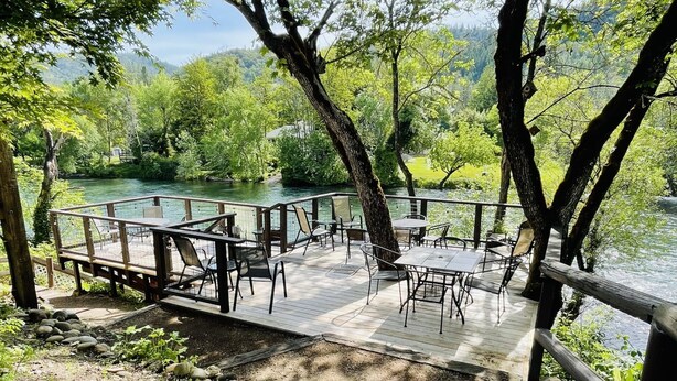 Una terraza de madera con mobiliario exterior, con vista a un río, rodeada de árboles y un paisaje natural.