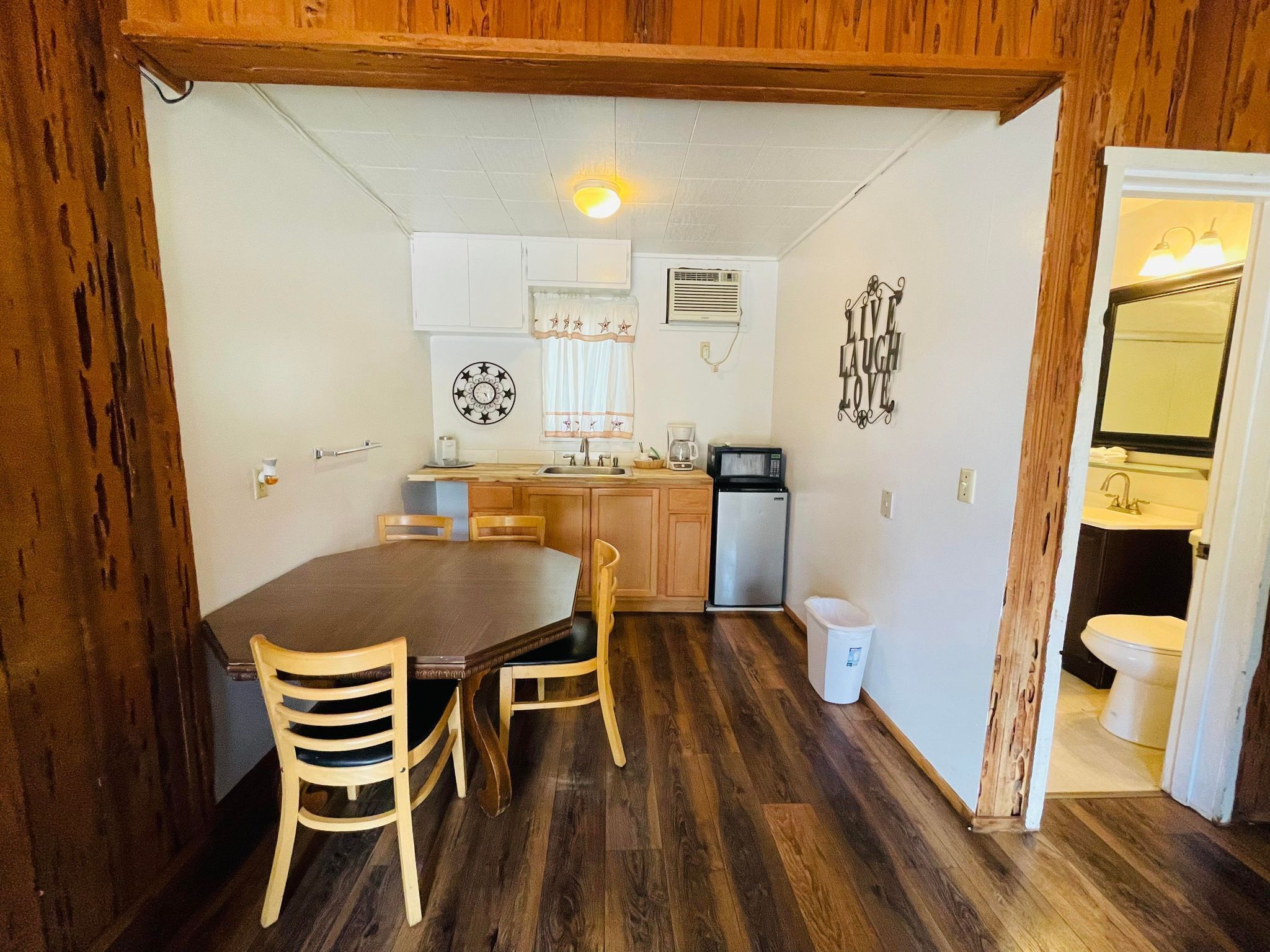 A compact kitchen with wooden cabinets, a refrigerator, a microwave, and a sink. There is a dining table with chairs and a clock on the wall.