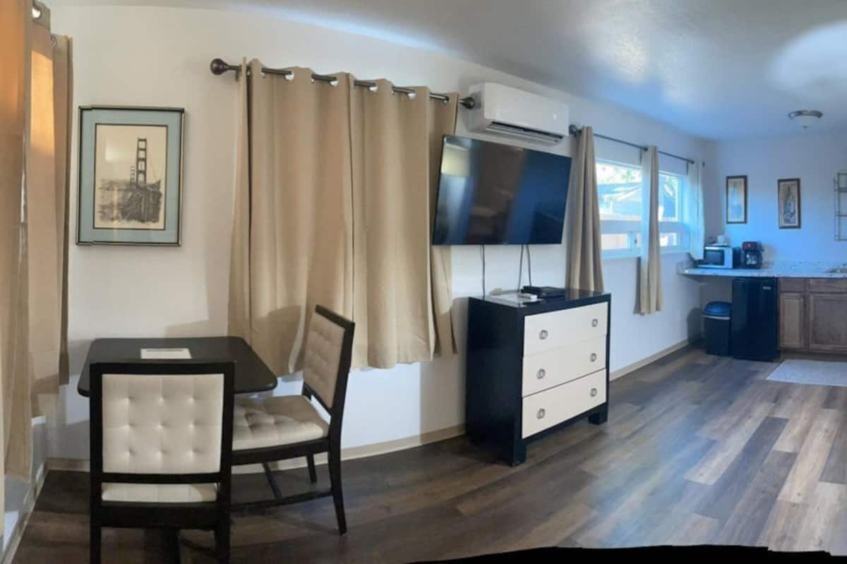 A living room with a sofa, a TV, a small table, and a chair. There is a kitchen in the background.