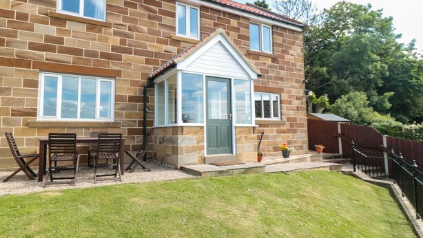A two-story house with a brick exterior, a front porch with a table and chairs, and a small garden area. - Cherry Garth (Whitby)
