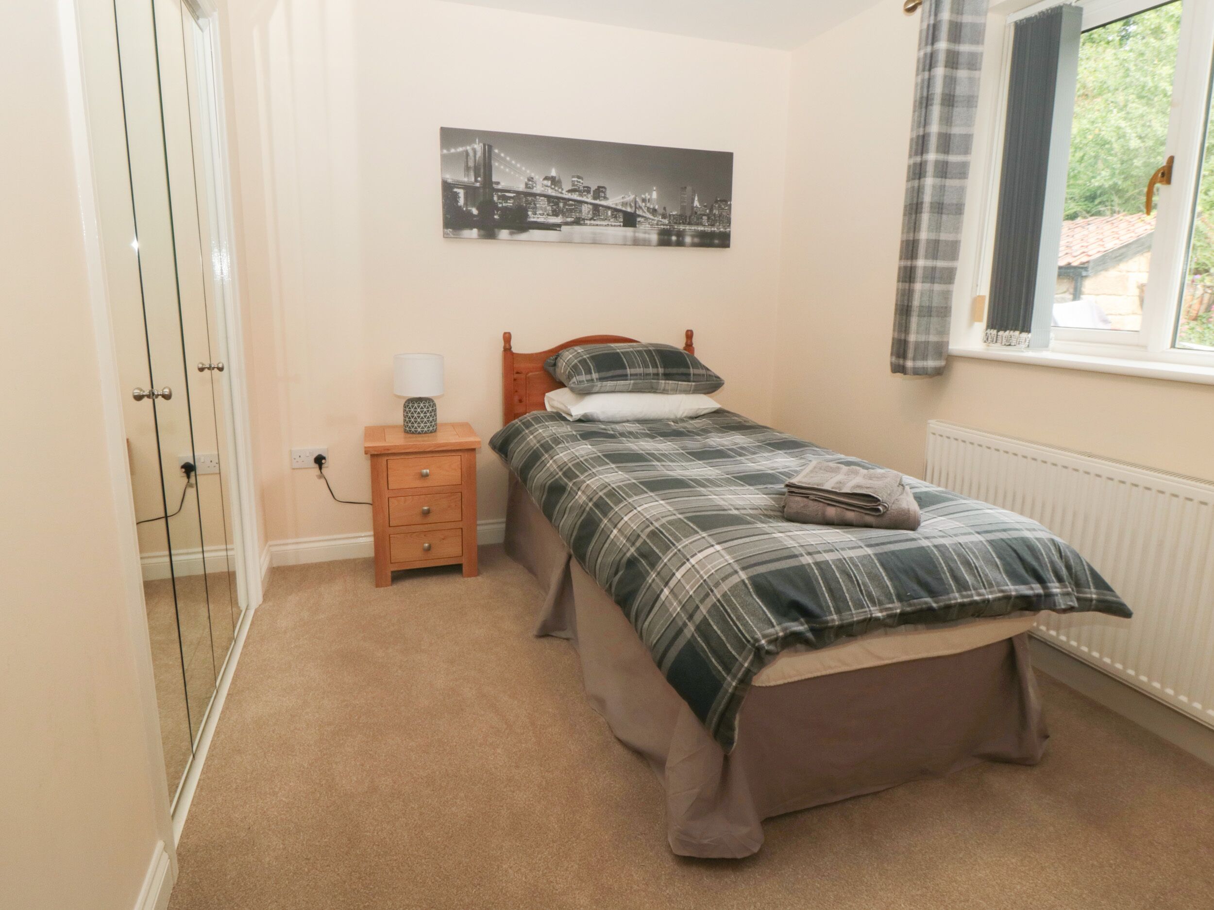 A bedroom with a bed, a nightstand, a lamp, a radiator, a window with curtains, and a cityscape painting on the wall.