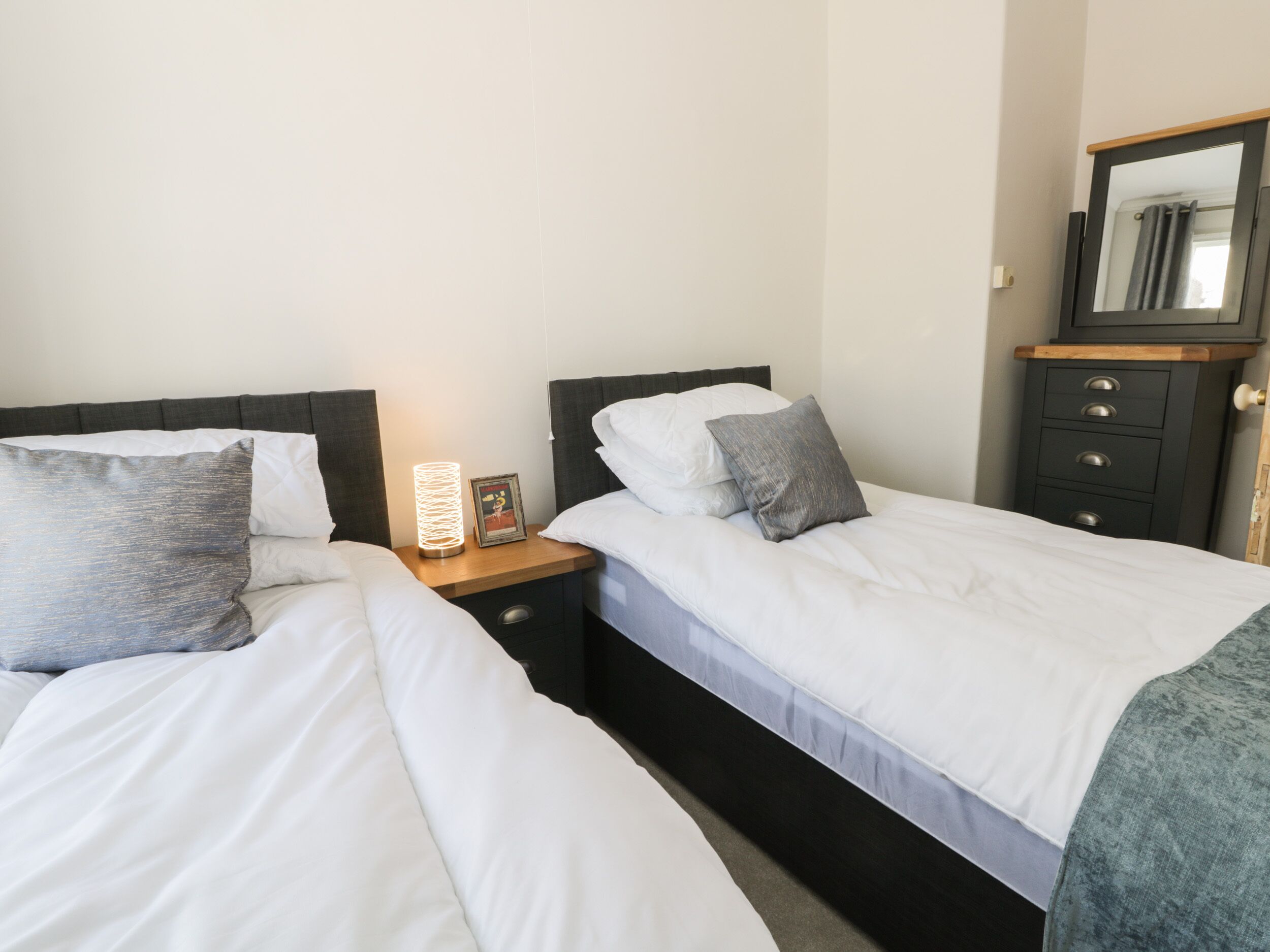 A bedroom with two single beds, a bedside table with a lamp, a dresser, and a mirror.