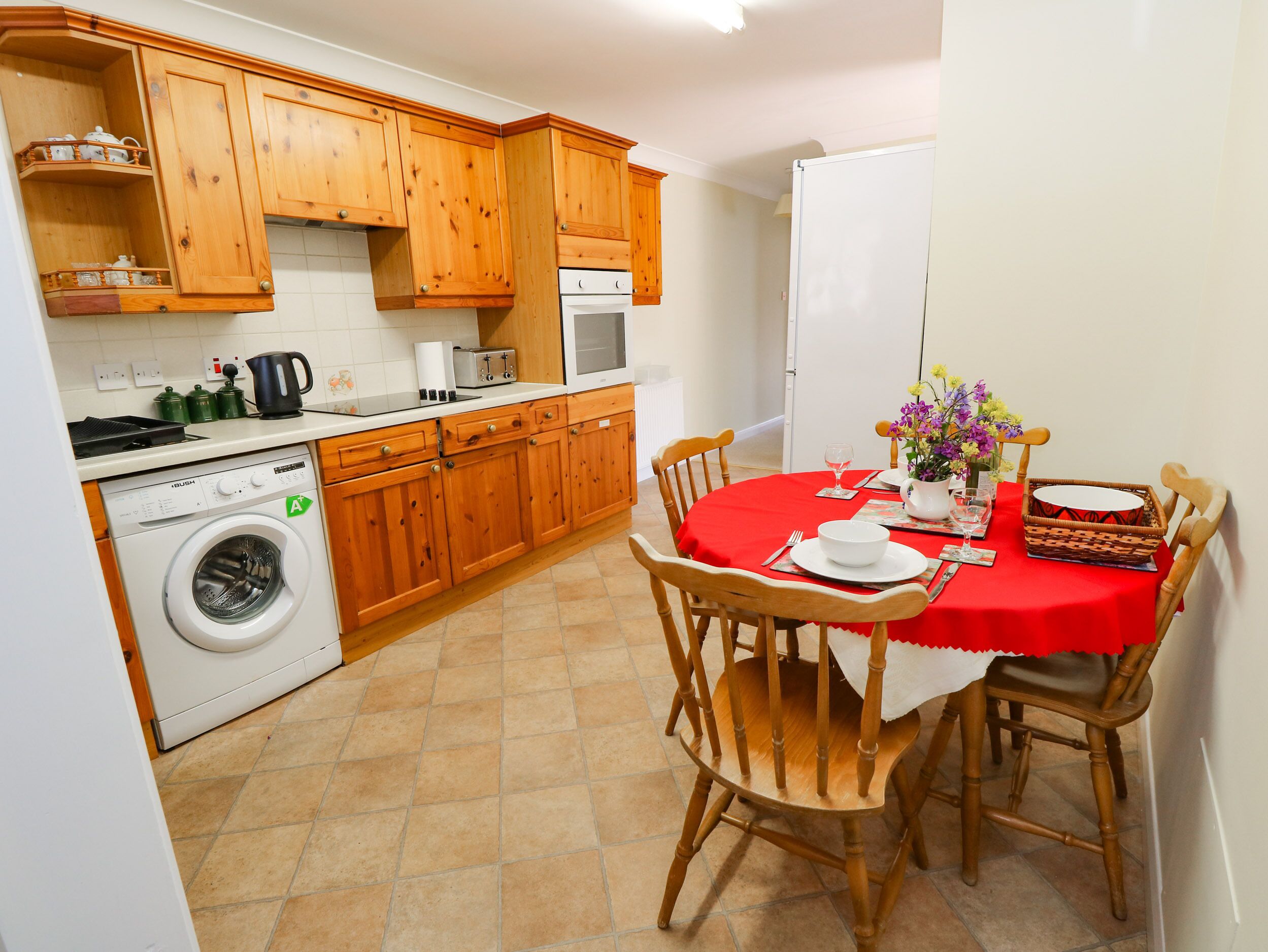 A compact kitchen with wooden cabinets, a washing machine, and a dining area with a red tablecloth.