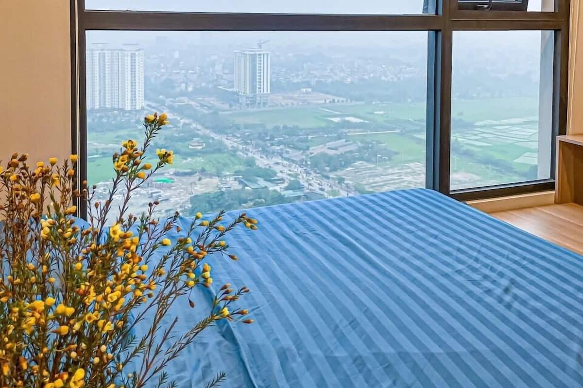 Une chambre avec une grande fenêtre, un lit recouvert d’une courtepointe à rayures bleues et un vase rempli de fleurs jaunes.