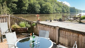 Ein runder Tisch mit Stühlen und Weinflaschen, mit Blick auf einen ruhigen See mit einer Holzterrasse und Kübelpflanzen.