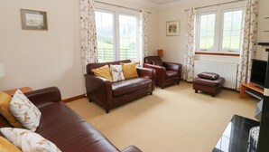 A living room with a brown leather sofa, a brown armchair, and a small ottoman. There is a television on a wooden stand to the right. - Brae Mhor Cottage (Fort William)