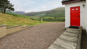 A building with a red door, gravel driveway, and a view of green hills in the background. - Brae Mhor Cottage (Fort William)