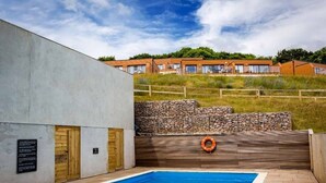 Una piscina con pared de piedra y cubierta de madera, rodeada por una valla y una colina cubierta de césped al fondo.