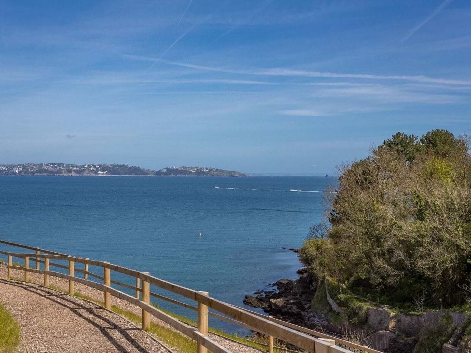 Udsigt over kysten med en træpromenade, der fører ned til havet, klippefyldt terræn og en fjerntliggende kystlinje med bygninger.
