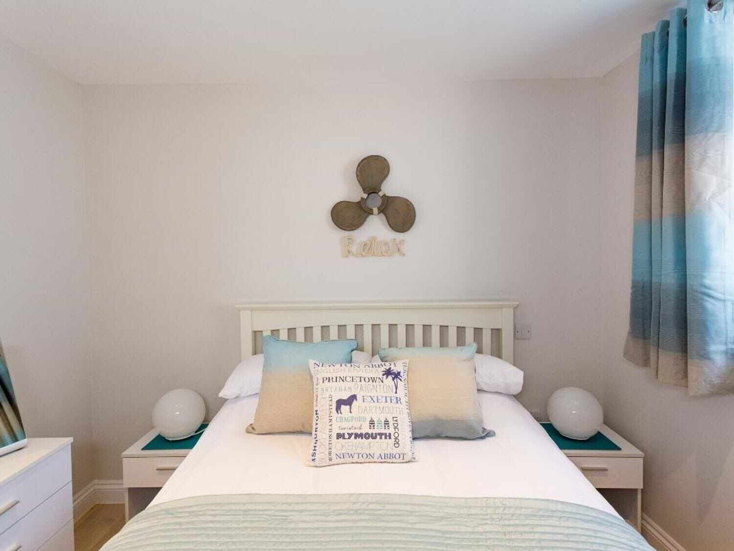 Un dormitorio con una cama blanca, dos mesitas de noche, un adorno en la pared y una cortina azul.