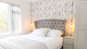 A bedroom with a large bed, two bedside tables, a headboard, and a window with blinds. - Harbour Place (Wells-next-the-Sea)