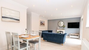 A modern living room with a dining area, a blue sofa, a fireplace, and a TV mounted on the wall. - Harbour Place (Wells-next-the-Sea)