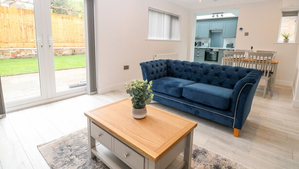 A modern living room with a blue sofa, a wooden coffee table, and a sliding glass door leading to a backyard. - Harbour Place (Wells-next-the-Sea)