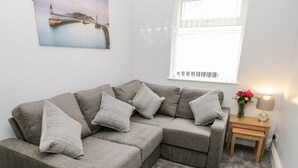 Um sofá modular cinza com várias almofadas, uma pequena mesa de madeira com uma luminária e flores, e um quadro de uma ponte na parede.
