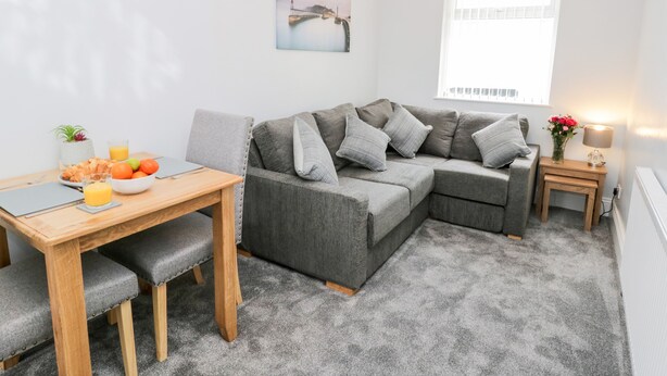 Uma sala de estar moderna com um sofá cinzento, uma mesa de jantar de madeira e uma pequena mesa lateral com uma luminária.