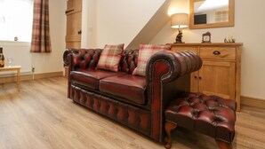 A cozy living room with a dark brown leather sofa, matching ottoman, and plaid cushions. A wooden cabinet with a mirror and clock is against the wall.