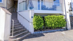 A two-story building with a staircase leading to a balcony, a glass railing, and a small garden area with green shrubs. - Ocean View Apartment (Colwyn Bay)