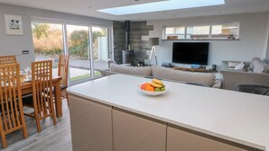 A modern kitchen with a central island, dining area, and a living space with a TV. - Dolwern (Newport)