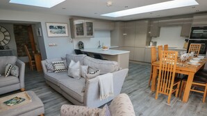 A modern living room with a grey sofa, patterned cushions, and a coffee table. A dining area with wooden chairs and a table set for a meal is visible. The kitchen has a minimalist design with grey cabinets and a built-in oven. - Dolwern (Newport)
