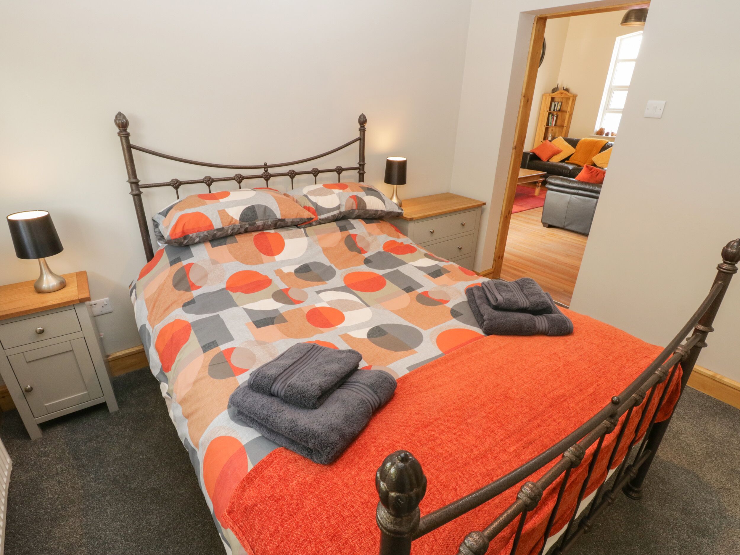 A neatly made bed with a patterned bedspread, two bedside lamps, a wooden nightstand, and a view into another room with a sofa and a wooden cabinet.