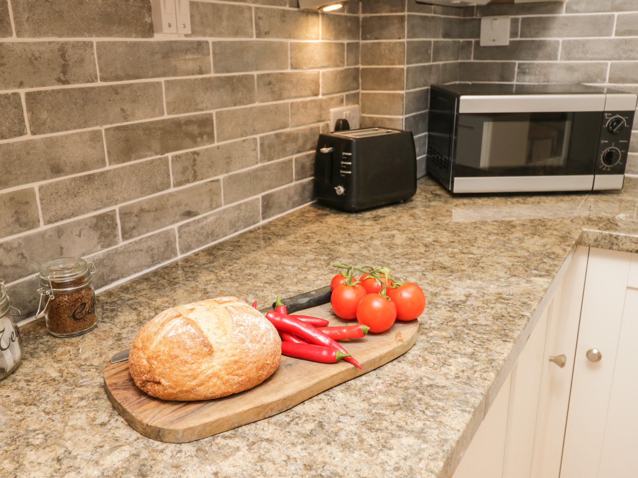 Une cuisine avec un plan de travail en granit, une miche de pain, des tomates et des piments rouges sur une planche à découper.