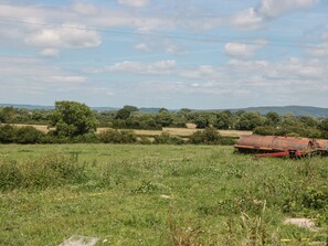 A rural landscape with a rusty metal structure in the foreground, surrounded by green fields and trees under a partly cloudy sky. - The Old Cow Shed (Banwell)