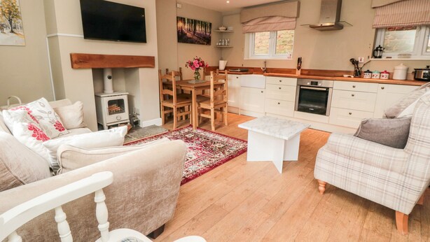 A cozy living room with a fireplace, a dining area with a table and chairs, and a kitchen with white cabinets and a window. - Swainby Cottage (Northallerton)