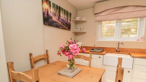 A dining area with a wooden table, chairs, a vase of pink flowers, and a painting of a forest scene. - Swainby Cottage (Northallerton)