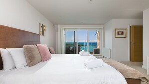 A large bed with white linens and multiple pillows, a wooden headboard, and a view of the sea through a sliding glass door. - Sandbanks (Penzance)