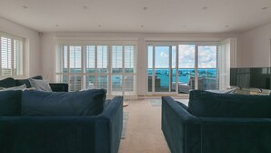A spacious living room with a large sliding glass door leading to a balcony with a view of the sea and boats. There are two dark-colored sofas and a flat-screen TV on a stand. - Sandbanks (Penzance)