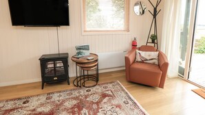 A living room with a flat-screen TV, a small table with a book, a sofa with a decorative pillow, a patterned rug, and a window with blinds. - 3 Valley View Lodges (York)