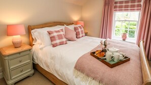 Ein ordentlich bezogenes Bett mit einer rosa karierten Tagesdecke, ein Nachttisch mit Lampe und ein Fenster mit Vorhängen.