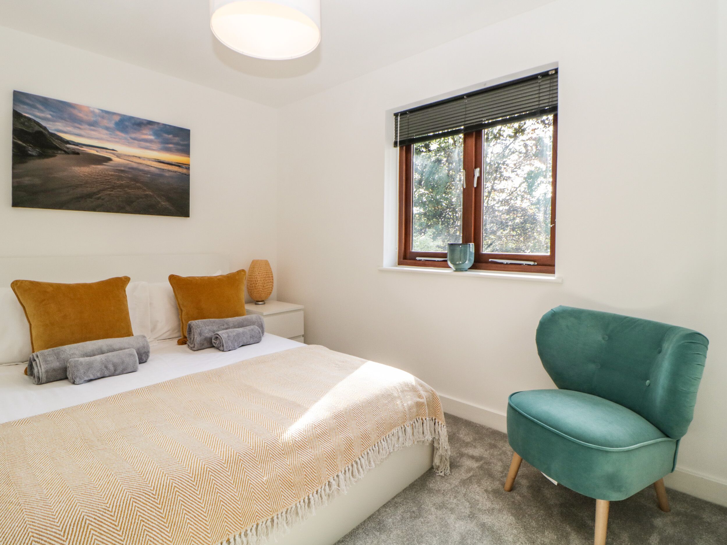 Een slaapkamer met een bed, een blauwgroene stoel, een raam met jaloezieën, een nachtkastje en een schilderij aan de muur.