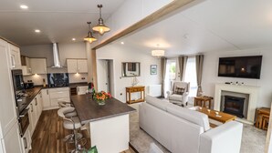 A modern kitchen with white cabinets, a wooden island, and bar stools. There is a living area with a sofa, coffee table, and fireplace. A TV is mounted on the wall. - Bramblewick (Saltburn-by-the-Sea)