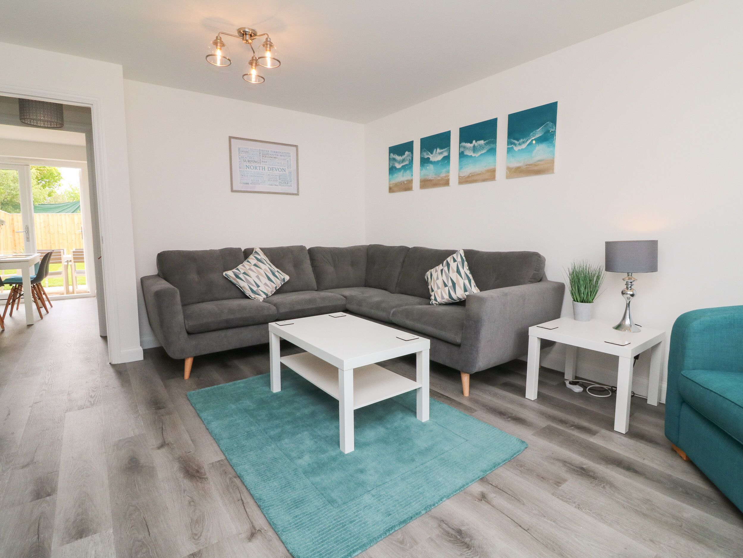 A modern living room with a grey sectional sofa, white coffee table, and a teal rug. There are three framed artworks on the wall and a small side table with a lamp.