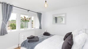 A bedroom with a bed, grey curtains, a window with flowers, a radiator, and a hanging light fixture. - Mischa Cottage (Bradford)