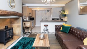 A modern living room with a fireplace, a sofa, a coffee table, and a kitchen in the background. - Mischa Cottage (Bradford)