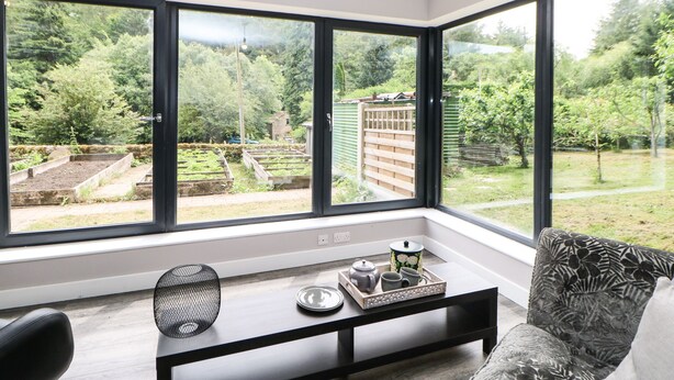 Une pièce d’angle avec de grandes fenêtres, un canapé noir, une petite table sur laquelle se trouve un plateau avec divers objets, et une vue sur un jardin et des arbres.